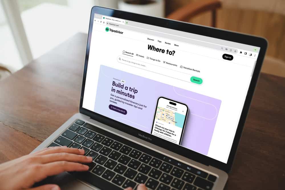 A person using a laptop displaying a travel planning website considers electronics waste disposal options during their trip. The screen showcases destinations, hotels, and attractions. A smartphone image features a map as hands type on the keyboard, mindful of the need to recycle computers responsibly.