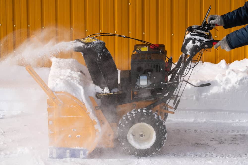 Against a yellow wall backdrop, a person operates an orange snowblower, effortlessly clearing snow. Clad in dark clothing and gloves, they grip the machine's handles with precision. It's as efficient as finding new ways to recycle laptops during E-waste disposal, ensuring a cleaner future.