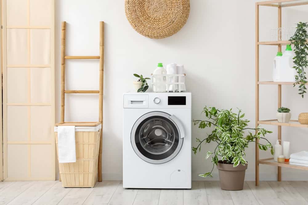 A modern laundry room with a white washing machine, a wicker laundry basket, and potted plants encourages sustainable living. Wooden shelves hold detergent bottles and folded towels. A round woven wall decor hangs above, while a wooden ladder leans against the wall, reminding one of creative E-waste disposal ideas.
