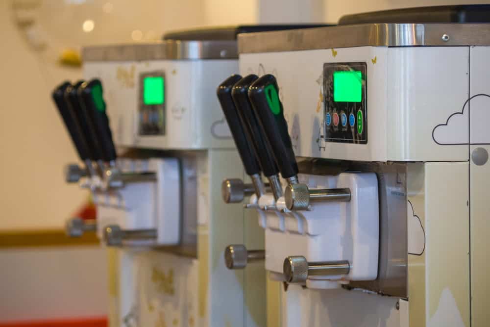 Close-up of two soft-serve ice cream machines with digital displays and black levers, showcasing a design inspired by clouds. Positioned on a countertop, these machines highlight the importance of sustainability, much like electronics recycling for a greener future.