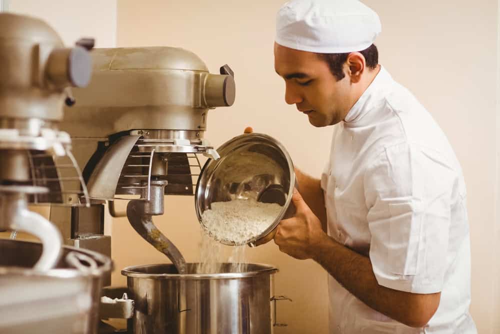 In a professional kitchen, a baker in a crisp white uniform and hat pours flour from a metal bowl into a large industrial mixer, reminiscent of the precision seen in electronics recycling where every component finds its perfect place.