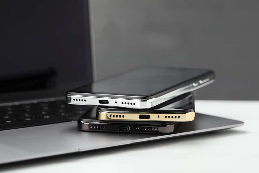 Three smartphones in silver, black, and gold are stacked on a slightly open laptop, highlighting the charging ports against a blurred background. This image underscores the importance of electronics recycling to manage waste and promote sustainability.