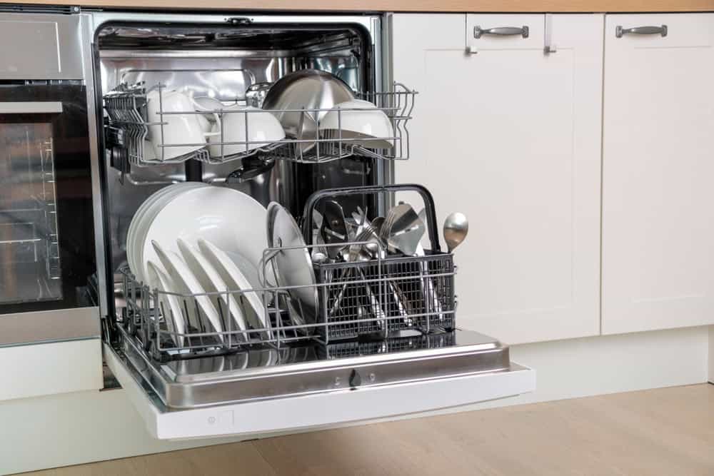 The open dishwasher showcases neatly arranged white plates, bowls, pots, and silverware. Set against a kitchen backdrop of white cabinetry with metallic handles, the scene emphasizes cleanliness. Remember to prioritize eco-friendly habits like electronics recycling in your home for a sustainable lifestyle.