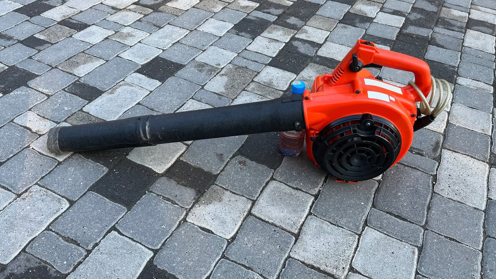 A red leaf blower rests on a pavement of interlocking gray and black bricks, like a reminder to recycle electronics responsibly. The blower's nozzle extends to the left while its handle is visible on the right.