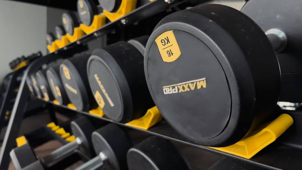 A close-up image of a rack filled with various black dumbbells labeled "MAXXPRO," each neatly arranged on yellow stands inside a gym. Just like the careful arrangement of electronics for recycling, the attention to detail here is impeccable, with one dumbbell visibly marked as 16 kg.