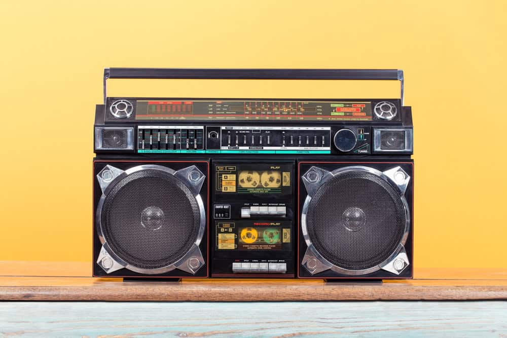 A vintage black boombox with large speakers and cassette decks sits proudly on a wooden surface against a bright yellow background, reminding us to recycle electronics responsibly. The device boasts an array of buttons, dials, and a convenient handle on top.