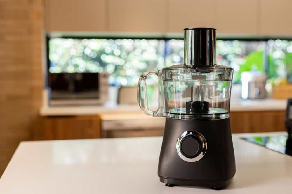 A black and silver food processor sits on a white kitchen counter. The background features blurred wooden cabinets, a microwave, and a window with greenery outside, reminding us of the growing need for electronics recycling to protect our environment.