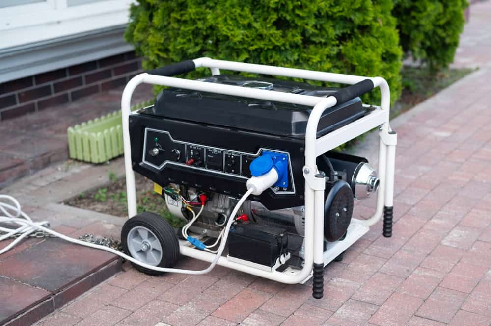 A portable generator with a black and white frame sits on a brick pathway, its power cable ready for action. Equipped with wheels for easy mobility, it's near a green bush, white wall, and a planter&mdash;a reminder to recycle computers and embrace sustainable electronics waste disposal.