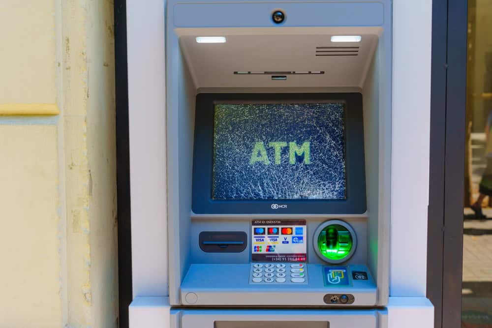 Close-up of an ATM machine with a cracked screen displaying the word "ATM" in large letters. The machine, reminiscent of obsolete electronics awaiting recycling, is built into a wall and features a keypad, card slot, and cash dispenser.