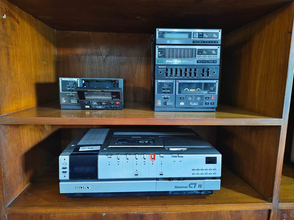 A wooden shelf showcasing vintage audio equipment, like a tape deck and stereo components. Below them sits a Betamax VCR labeled "Sony C7." These 1980s devices, with metallic and black finishes, remind us of the importance of electronics waste disposal to preserve such nostalgic treasures.