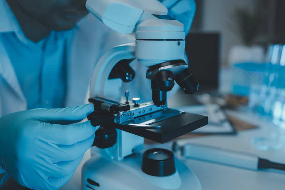 A scientist wearing blue gloves carefully adjusts an electronics recycling sample on a microscope stage in the lab. The image is illuminated with a bluish hue, while blurred laboratory equipment fills the background, emphasizing a commitment to innovative e-waste disposal solutions.