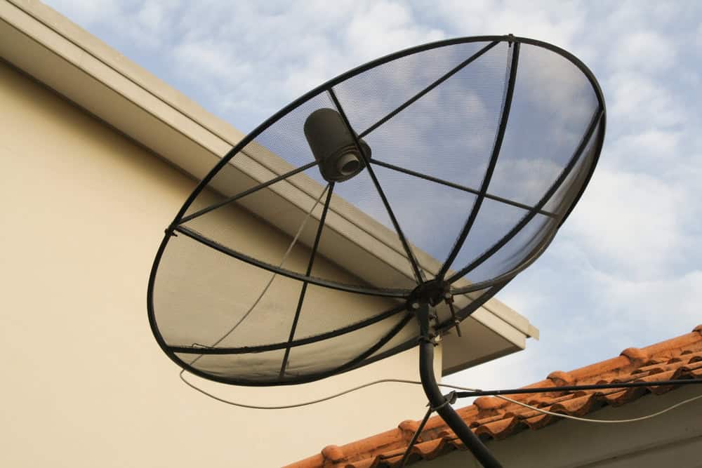 A large satellite dish faces upward, mounted on a building with white and beige walls. Part of a red-tiled roof is visible below the dish. The scene subtly echoes the theme of electronics recycling amidst a sky that's blue with light clouds.
