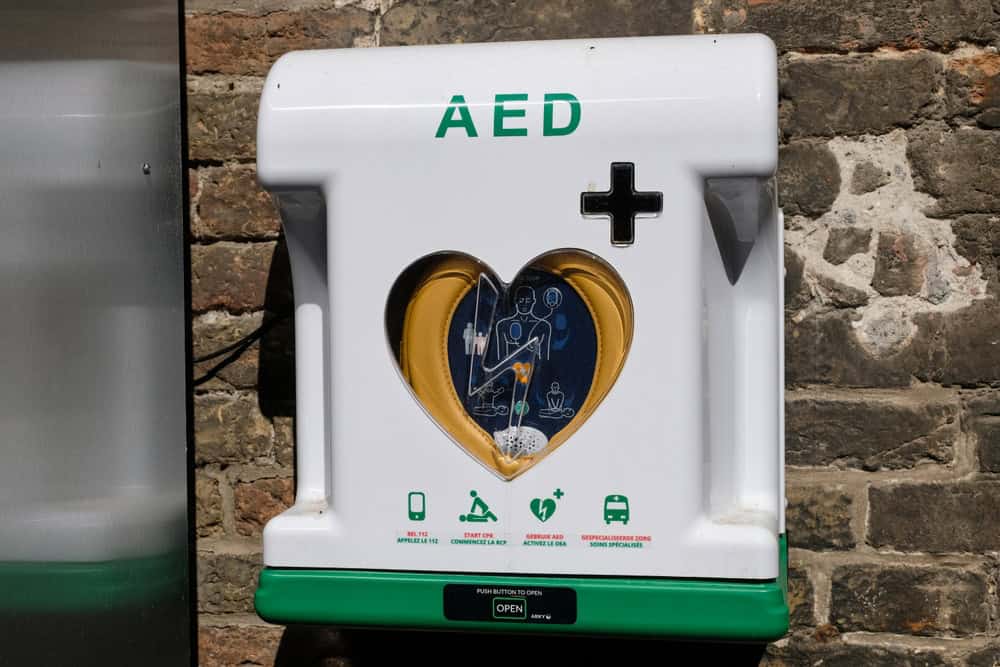 AED device mounted on a brick wall, with a heart-shaped transparent center revealing a blue and gold interior crafted from recycled electronics. The panel includes instructions, symbols for use, and a green "open" tab at the bottom.