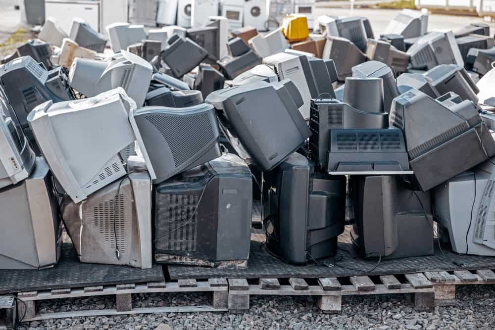 A large pile of discarded CRT televisions and monitors on wooden pallets, surrounded by gravel. The electronics, various shades of gray and black, are stacked haphazardly outdoors, highlighting the need for proper e-waste disposal through electronics recycling efforts.