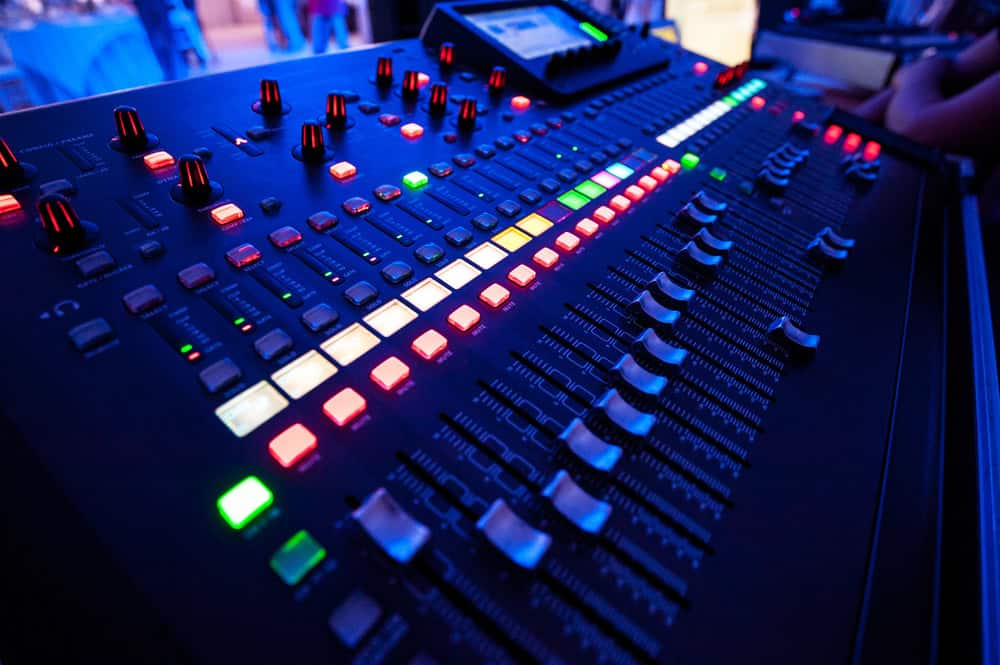 Close-up of an audio mixing console with glowing multicolored buttons and sliders, showcasing how obsolete electronics can be repurposed in creative ways. The console is illuminated under dim lighting, creating a vibrant atmosphere. Some knobs and screens are visible in the background.
