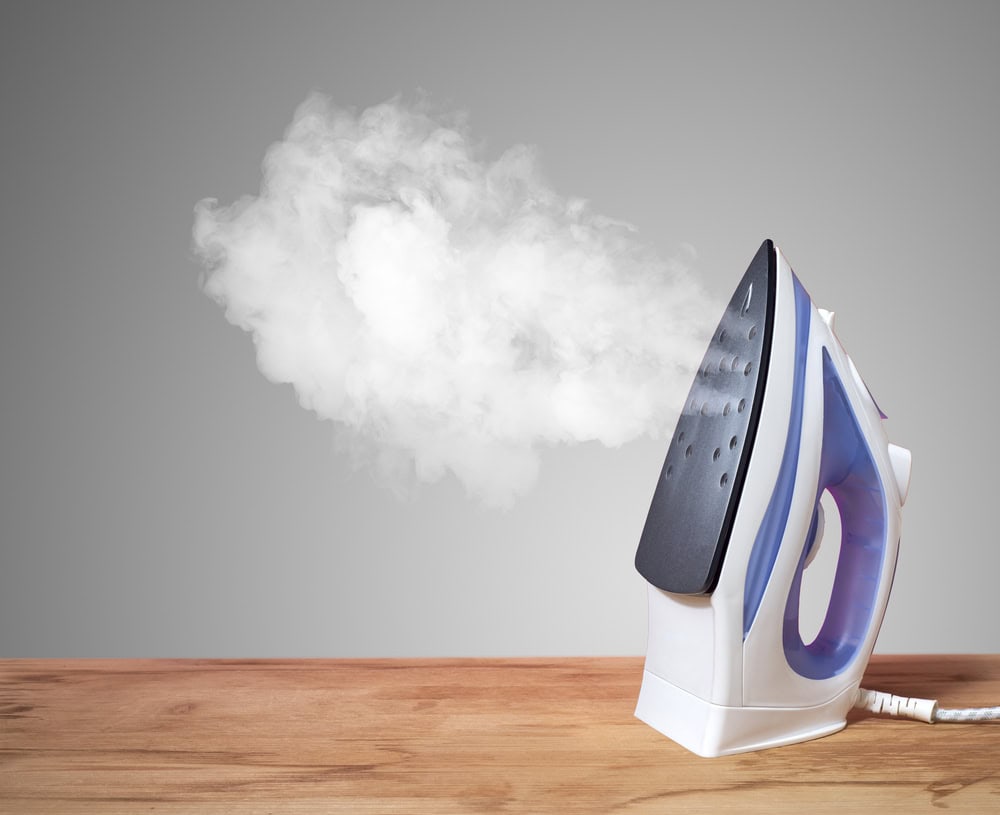 A steam iron releasing a cloud of white steam, placed upright on a wooden surface against a gray background. The mostly white iron with blue accents and a visible power cord serves as an important reminder of responsible electronics recycling.