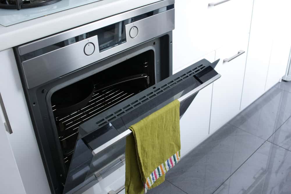 Open stainless steel oven with a partially pulled-out rack in a modern kitchen. A green towel with a colorful striped edge hangs from the oven door handle. The eco-friendly space, promoting electronics recycling, features white cabinets and gray tile flooring.