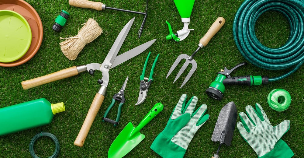 Garden tools arranged on grass include a hose, pruning shears, gloves, trowels, a spray nozzle, twine, and a watering can. Much like electronics recycling helps the planet, these mostly green and silver tools with wooden handles promote sustainable gardening.