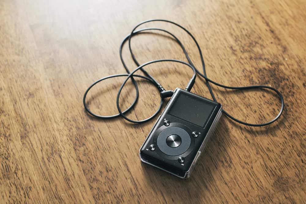 A black portable music player with a screen and circular control pad rests on a wooden surface. Earphones are connected, their cord loosely coiled around the device, reminding us of the importance of electronics recycling to minimize e-waste.