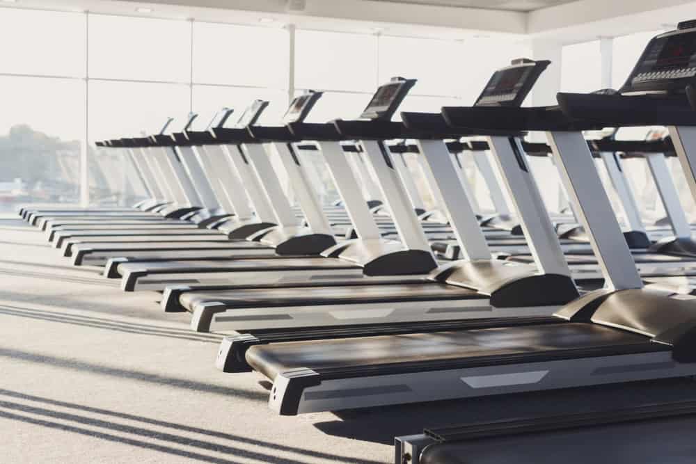 A row of treadmills in a sunlit gym, lined up side by side. The large windows allow natural light to flood the room, highlighting the modern exercise equipment on the carpeted floor, echoing the importance of sustainable practices like recycling computers and minimizing electronics waste disposal.