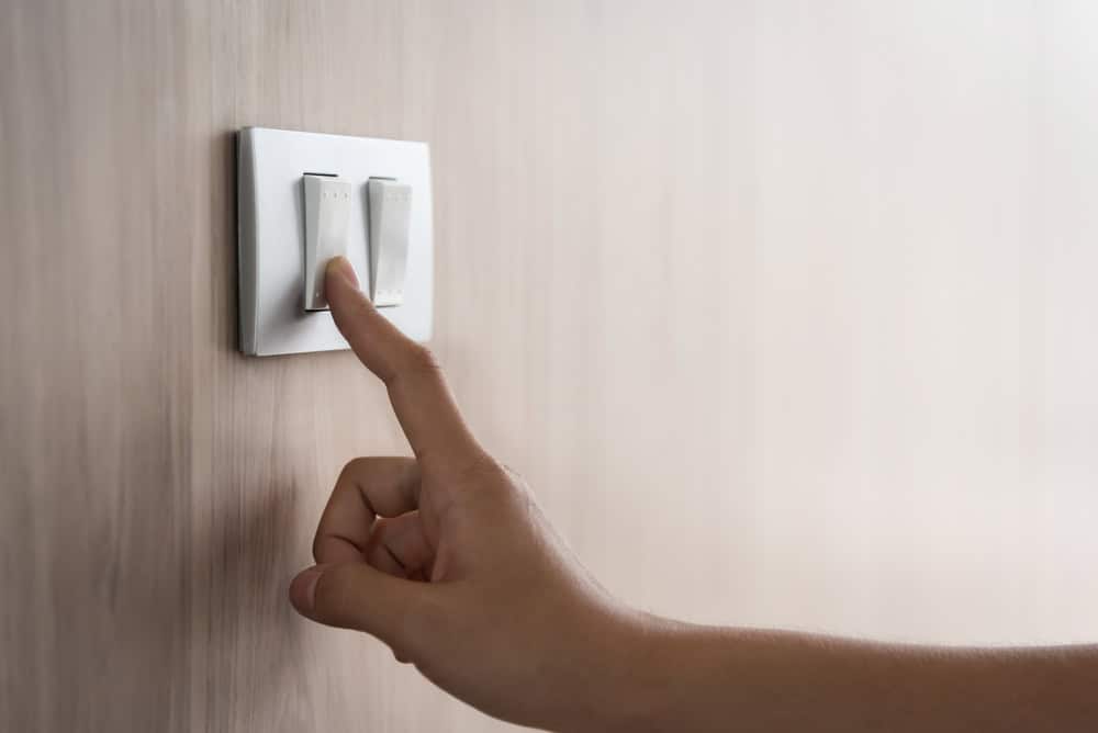 A hand is reaching towards a light switch on a wall, preparing to flick it. The wall has a smooth, wooden texture, and the switch plate is a simple, white rectangular design with two switches&mdash;perhaps made more sustainable through electronics recycling practices.