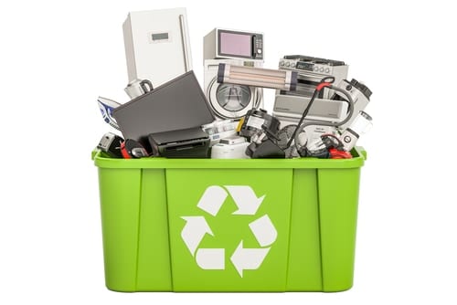 A green recycling bin overflowing with electronic waste, including appliances like a refrigerator, microwave, and speakers. This scene highlights the urgent need for proper electronics waste disposal. The bin is marked with a white recycling symbol, underscoring responsible E-waste disposal practices.