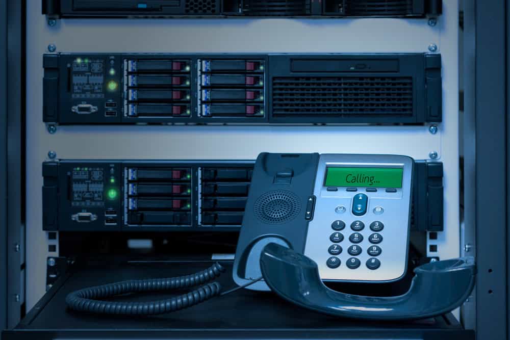 A digital office phone with a detached handset rests on a server rack, its display reading "Calling...". Behind it, multiple servers with blinking lights emphasize the need for responsible electronics waste disposal in this technology-driven environment.