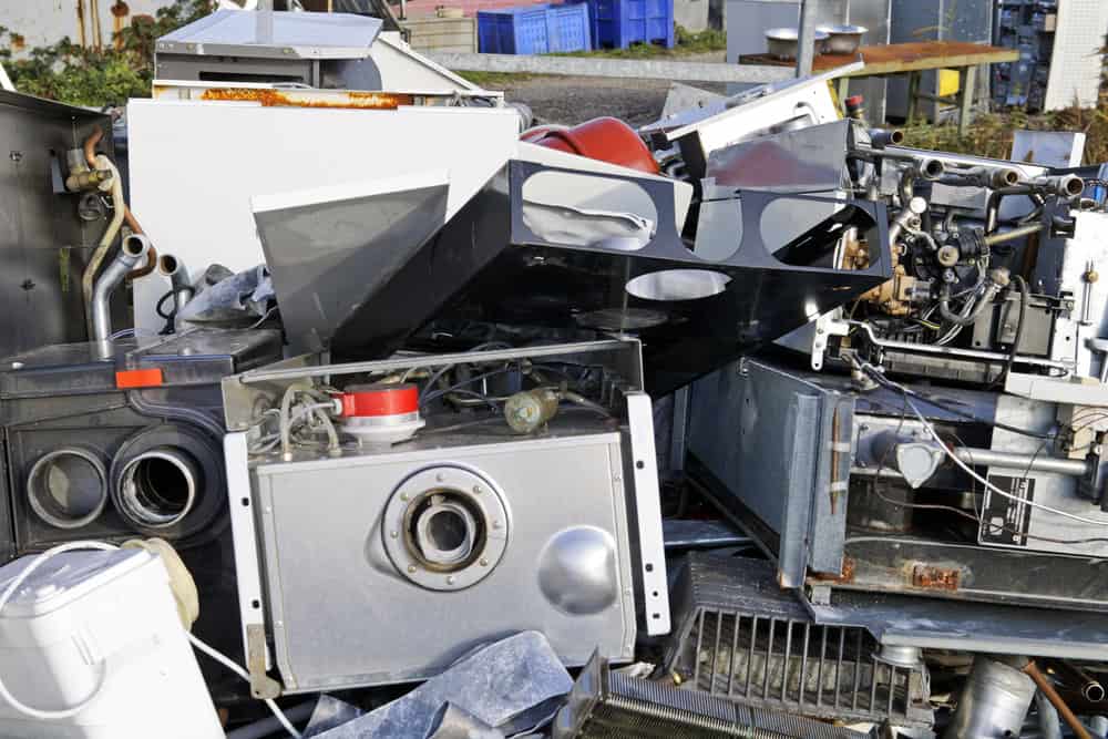 A pile of discarded electronic appliances and metal parts, including an oven, cables, and various machinery components, arranged messily outdoors highlights the urgency of electronics recycling. In the background, stacked blue bins and greenery underscore the need for responsible E-waste disposal.