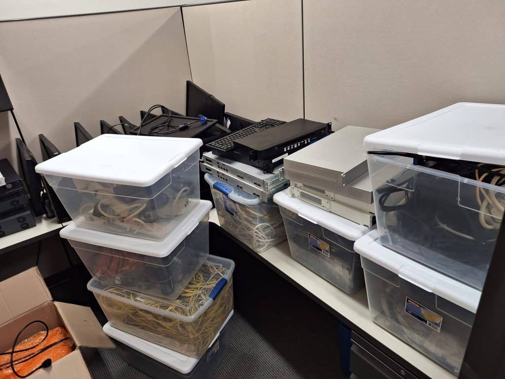 Stacks of clear plastic bins filled with network cables and electronic equipment await E-waste disposal in an office corner. A laptop and additional devices rest on top, surrounded by a few cardboard boxes and power cords, all part of the electronics recycling process.