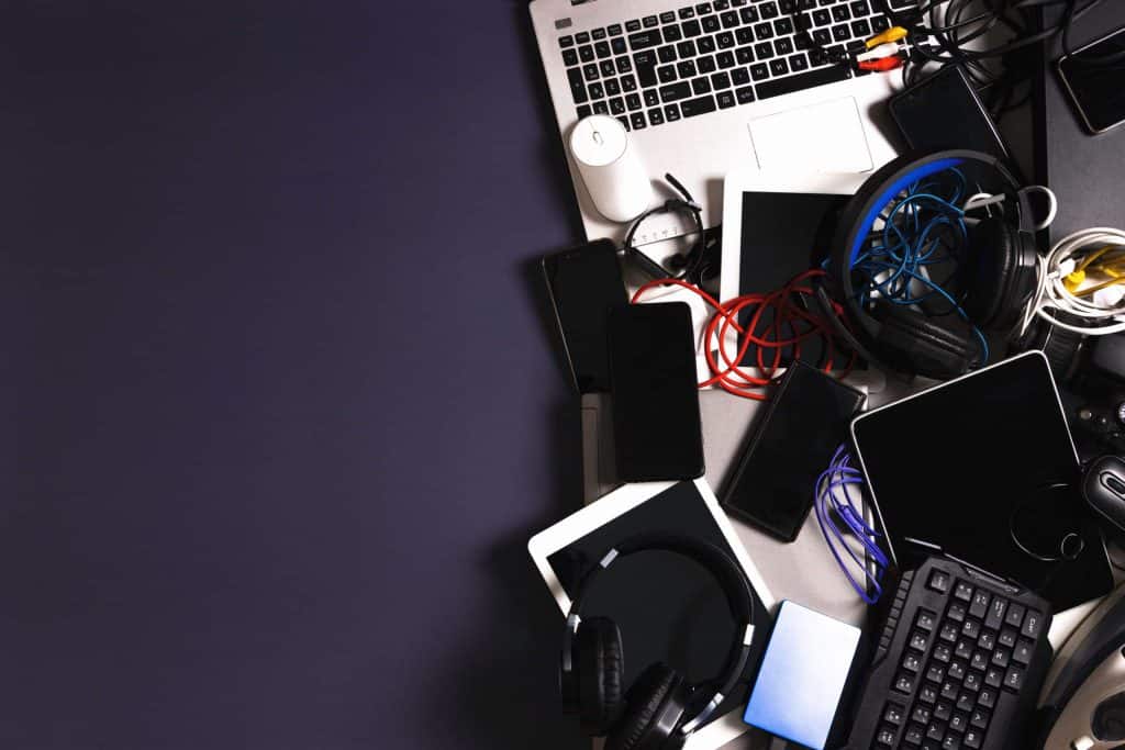 A cluttered workspace with various electronic gadgets, including a laptop, smartphones, tablets, headphones, and cables on a dark surface awaits electronics recycling. The left side is empty, creating a stark contrast with the busy right side brimming with tech in need of recycle laptops efforts.