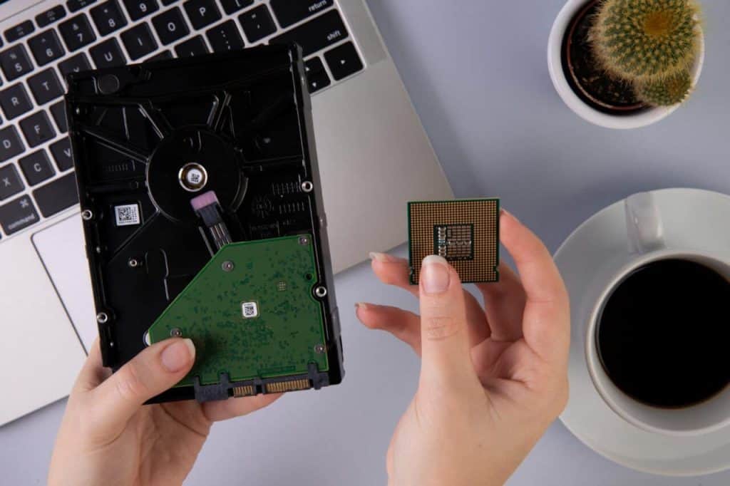 A person holds a computer processor and a hard drive above a laptop keyboard, contemplating electronics recycling. A cup of coffee and a small cactus in a pot are nearby on the desk, creating an environment that inspires mindful E-waste disposal.