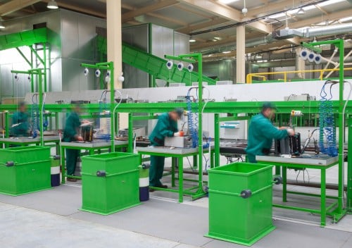 Workers in green uniforms are operating on an assembly line in a factory. They are handling equipment at workstations with bright green fixtures, part of a recycle laptops initiative. The background features industrial machinery and fluorescent lighting.