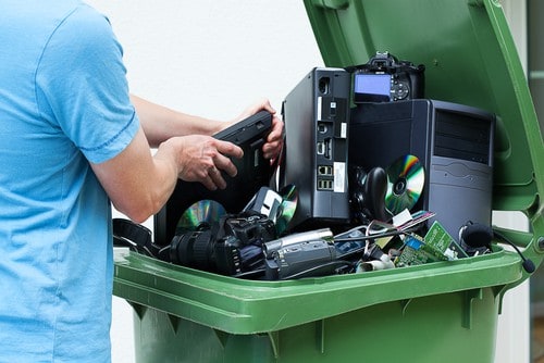 A person in a blue shirt places an old laptop into a green bin dedicated to e-waste disposal, filled with electronic waste like a desktop computer, camera, headphones, CDs, and cables.