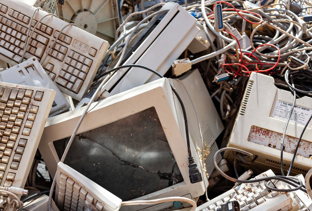 A pile of discarded electronic waste, including broken computer monitors, keyboards, and tangled cables, awaits electronics recycling in an outdoor setting.
