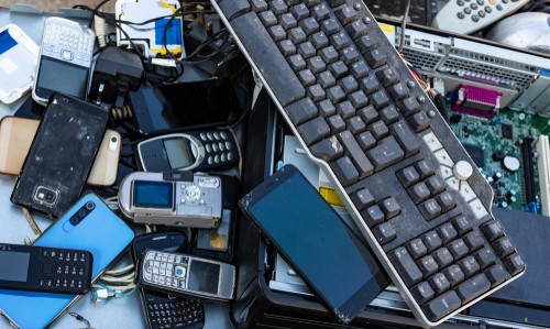 A pile of assorted electronic waste, including a keyboard, various mobile phones, a digital camera, and computer parts, awaits e-waste disposal. The visibly old and worn devices showcase outdated technology ready for recycling.