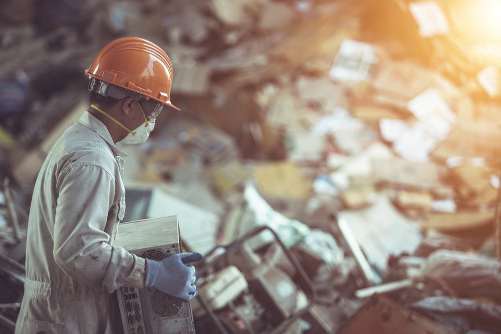 In a cluttered scrapyard, a worker in a hard hat, mask, and gloves examines an electrical device amidst various discarded items. Sunlight filters in from the corner, casting a warm glow over the scene of electronics recycling and efforts to recycle laptops.