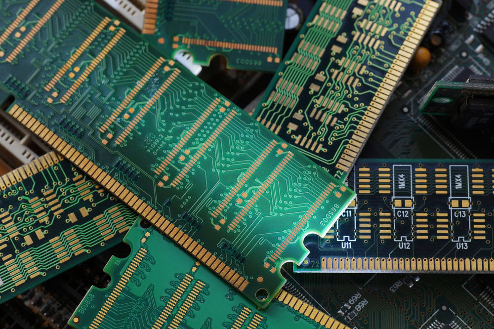 A close-up of a pile of green circuit boards shows intricate patterns of pathways and gold connectors, highlighting the importance of electronics waste disposal. The boards, from recycled laptops, are various sizes and overlap each other, with visible chips and electronic components.
