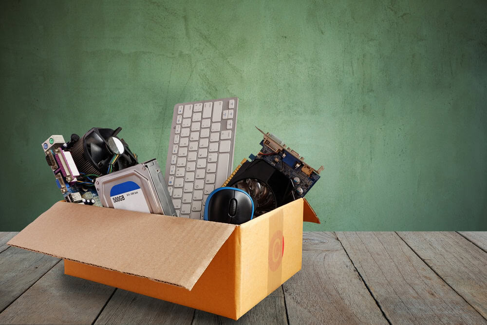A cardboard box on a wooden floor holds various computer components for e-waste disposal, including a keyboard, graphics card, hard drive, and mouse. The plain green wall in the background adds contrast to this recycling effort.