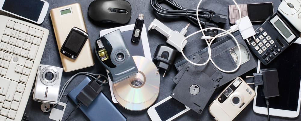 A cluttered assortment of outdated electronics and accessories, including smartphones, a computer mouse, cables, a VHS tape, a CD, a keyboard, floppy disks, and various chargers—perfect candidates for proper e-waste disposal—are scattered on a dark surface.
