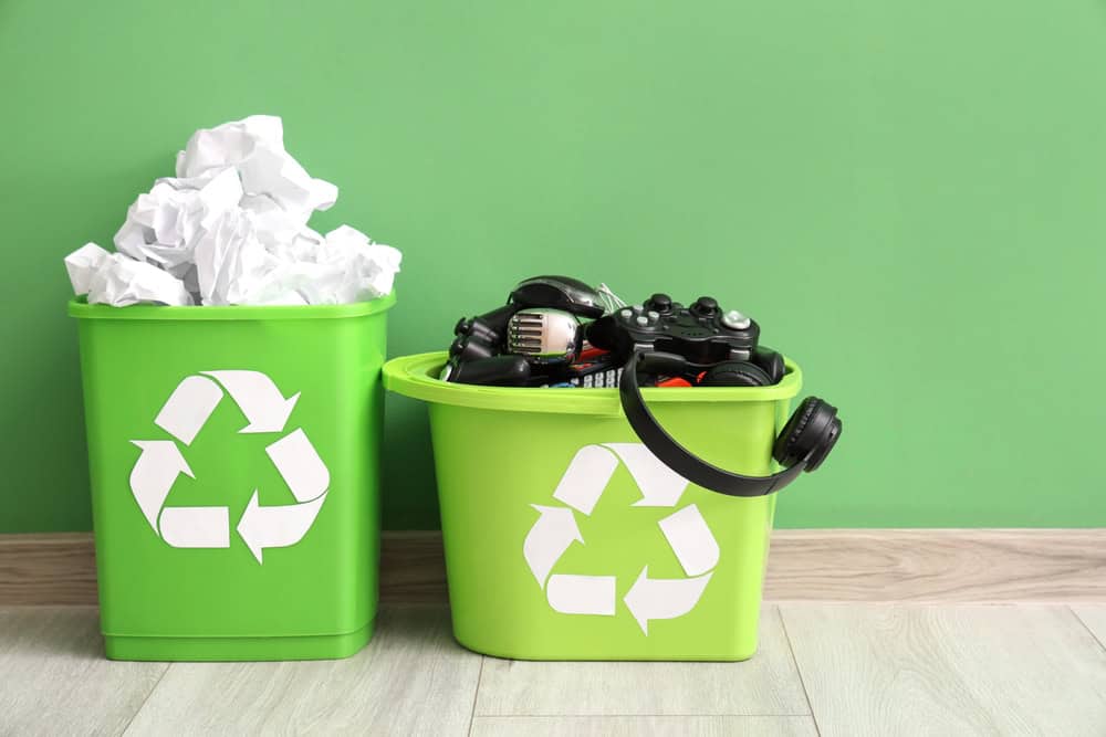 Two green recycling bins stand against a green wall on a wooden floor. The left bin overflows with crumpled white paper, while the right bin is dedicated to electronics recycling, holding items like headphones, gaming controllers, and a remote control. Both bins feature recycling symbols.