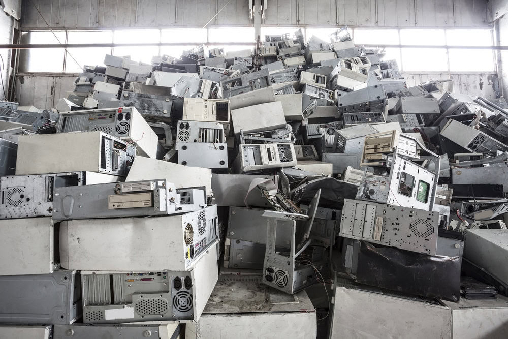 A large pile of discarded computer cases and components is stacked high in a warehouse, hinting at electronics waste disposal or recycling. The cases vary in size and color, with visible ventilations and sockets. Daylight filters through the windows, illuminating this hub for recycling computers.