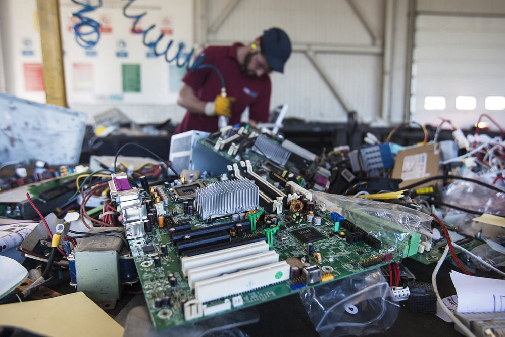 A worker in a cap and gloves skillfully manages electronics waste disposal at a recycling facility. In the foreground, various circuit boards and electronic components, including parts from recycled laptops, are scattered on a table.