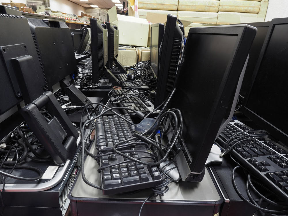 A cluttered pile of desktop computer monitors, keyboards, and tangled cables sits on a table, illustrating an electronics recycling scene with a disorganized setup. It's a vivid reminder of the importance to recycle computers responsibly.