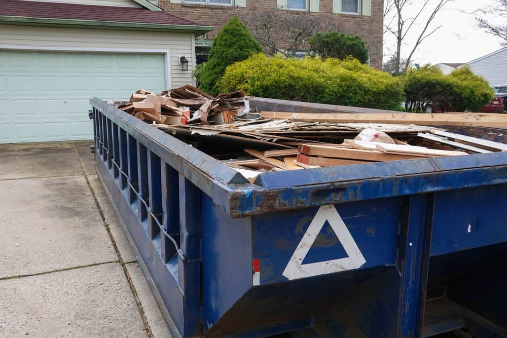 A large blue dumpster is filled with assorted debris, including wood and cardboard, placed on a driveway in front of a garage. Green shrubs and part of a house are visible in the background.