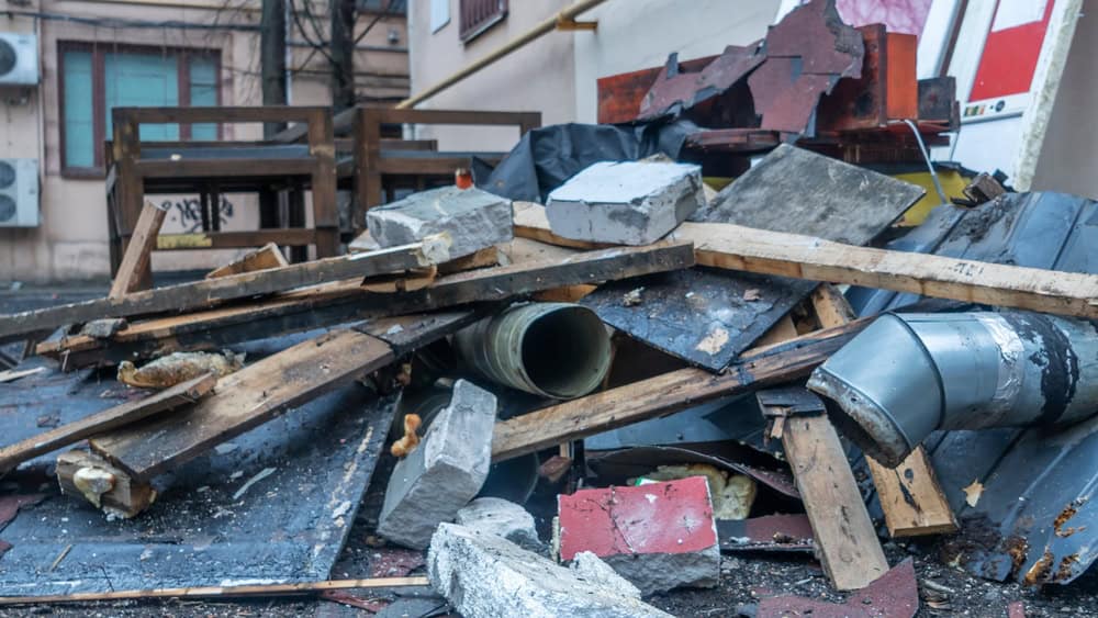 A pile of debris including wooden planks, concrete blocks, metal pipes, and broken boards is stacked haphazardly in an outdoor area near a building. The background shows part of the building with windows and an air conditioning unit.