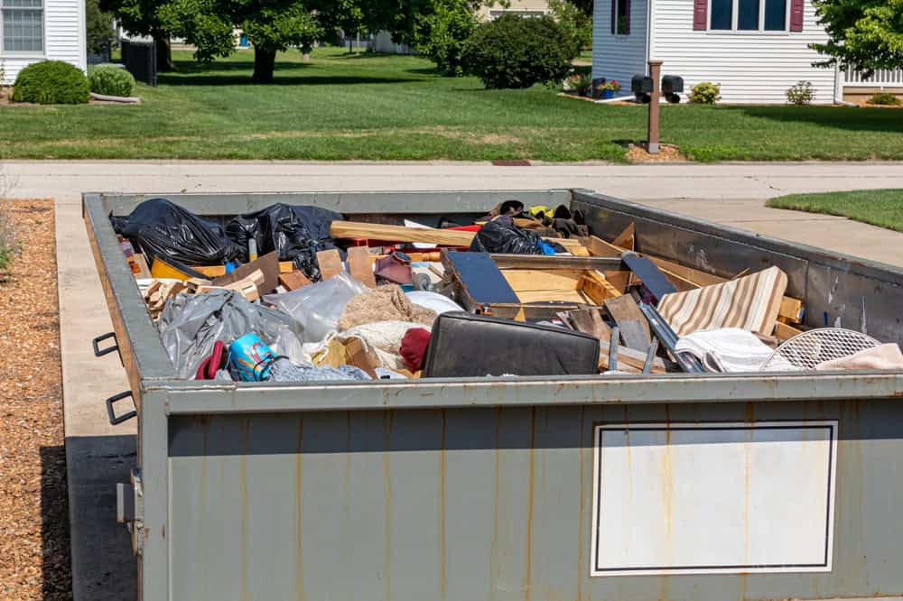 A large metal dumpster filled with various discarded household items, including wood, furniture, and black garbage bags. It's situated on a driveway in a suburban neighborhood with grass lawns and houses in the background.