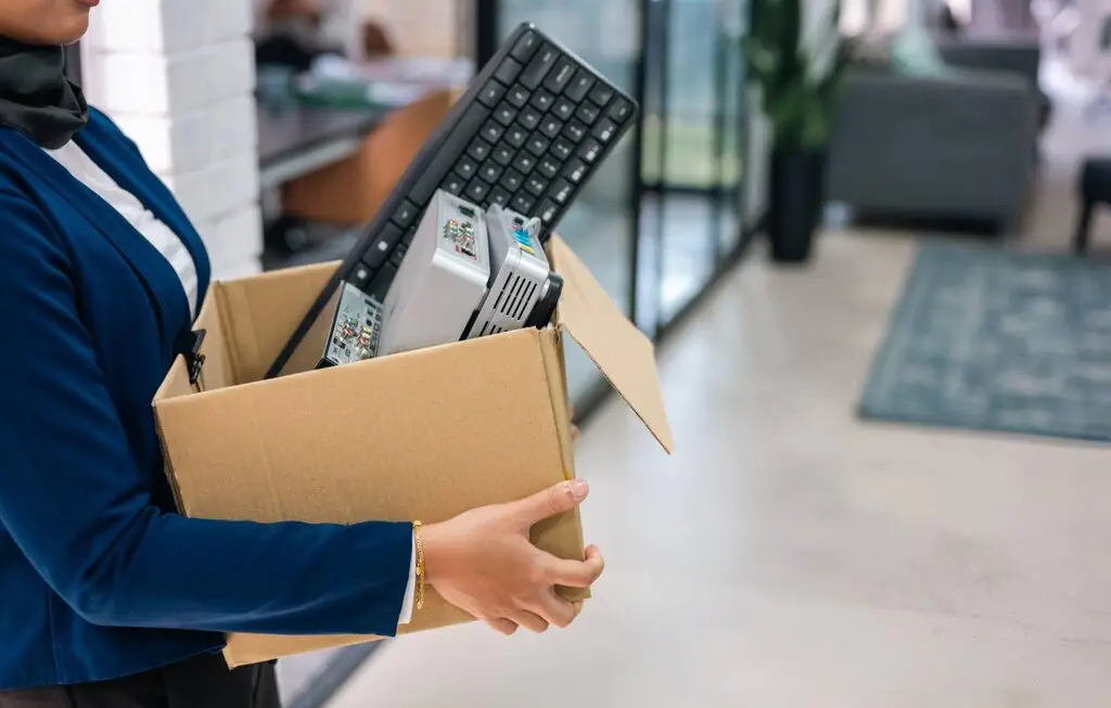 Person carrying a box filled with e-waste, including a keyboard and electronic components, in a modern office setting, highlighting sustainable electronics disposal and recycling efforts.
