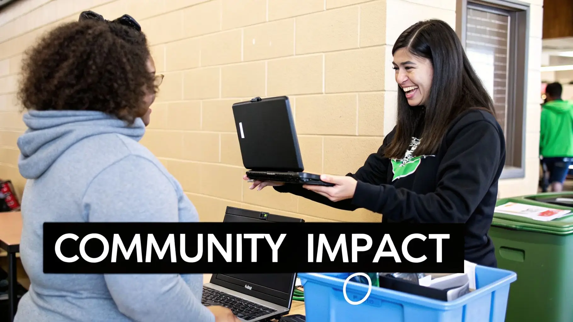 Two smiling women, one presenting a laptop to the other in a community setting.