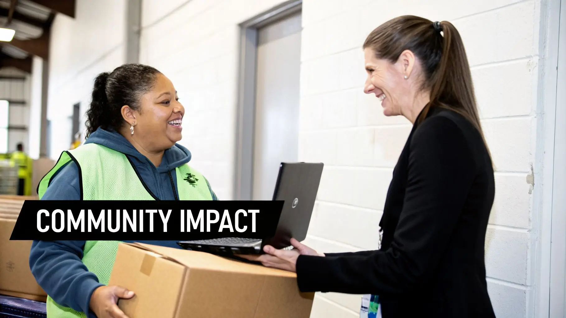 Two smiling women in a warehouse, one with a box and the other with a laptop, interacting.