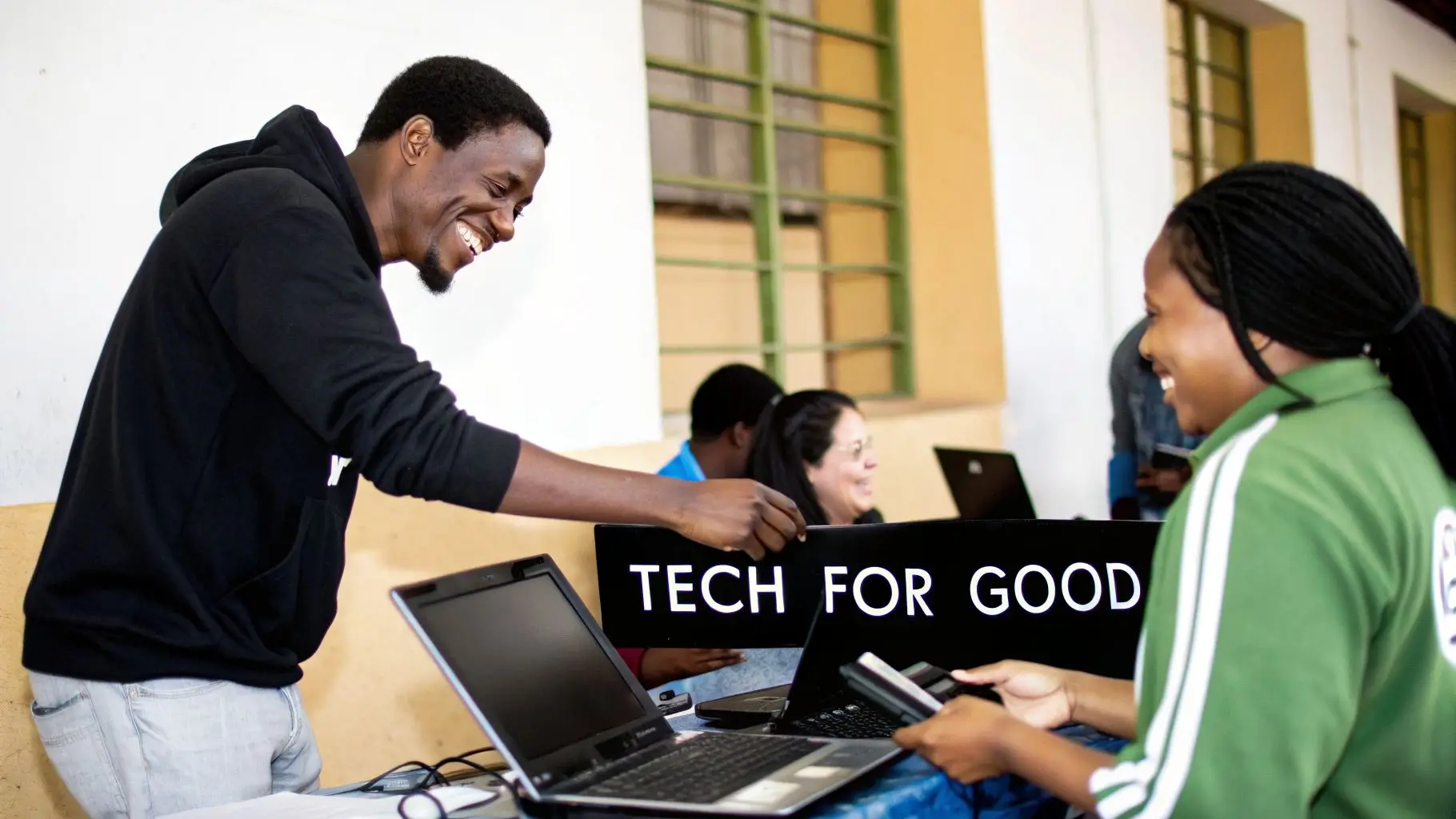A smiling man and woman interact with laptops and a 'TECH FOR GOOD' sign at an event.